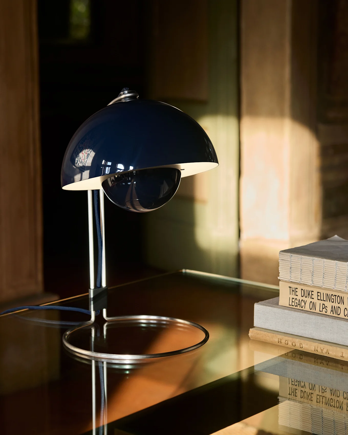 En mörkblå, klockformad bordslampa med krombas står på ett reflekterande bord bredvid en hög böcker.