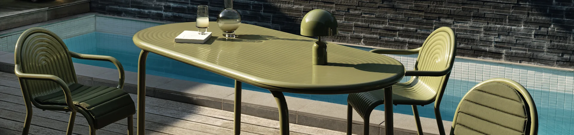 Olivgrön utomhusmatgrupp vid poolen med tre stolar, ett ovalt bord, en bok, en dryck, en karaff och en lampa.