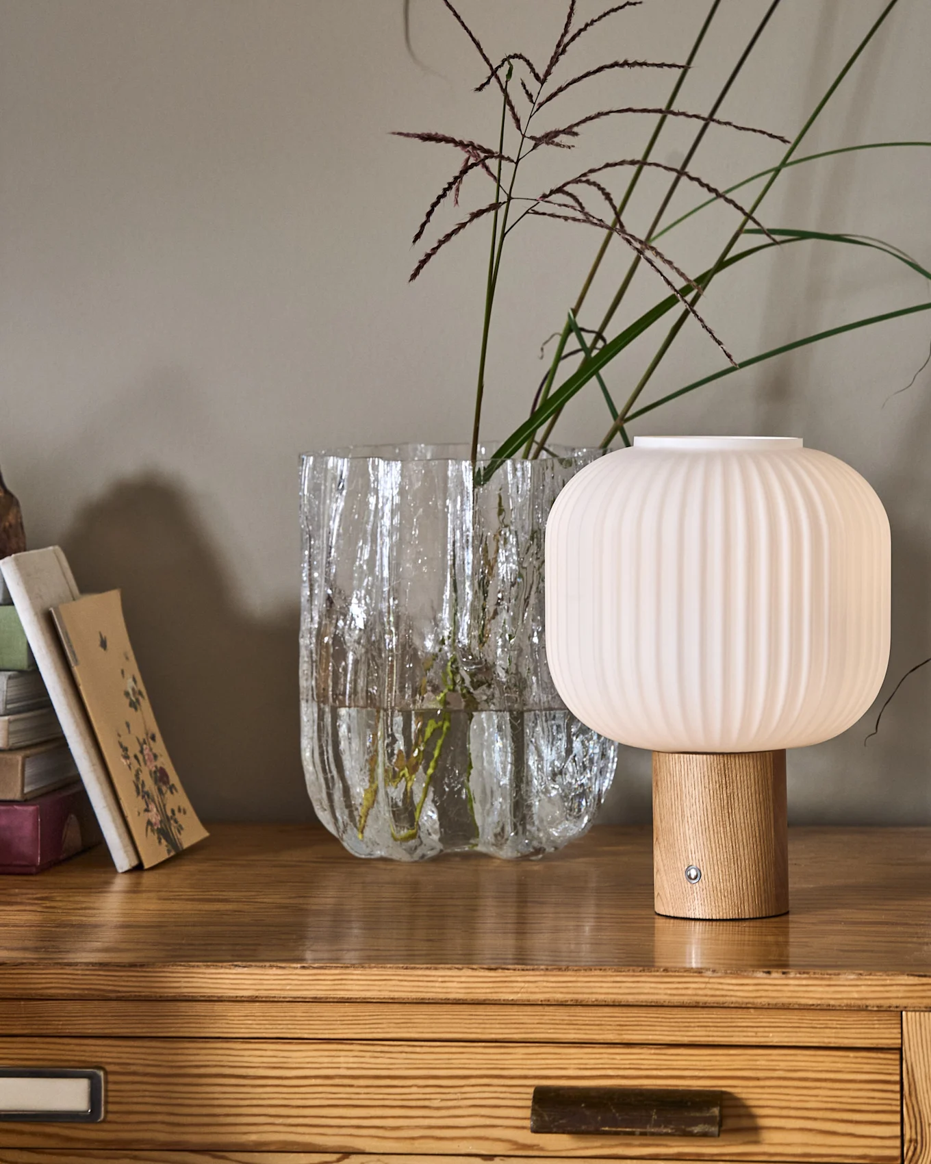 En räfflad, svampformad lampa med träfot bredvid en texturerad glasvas med växter och en hög böcker på en byrå i trä.