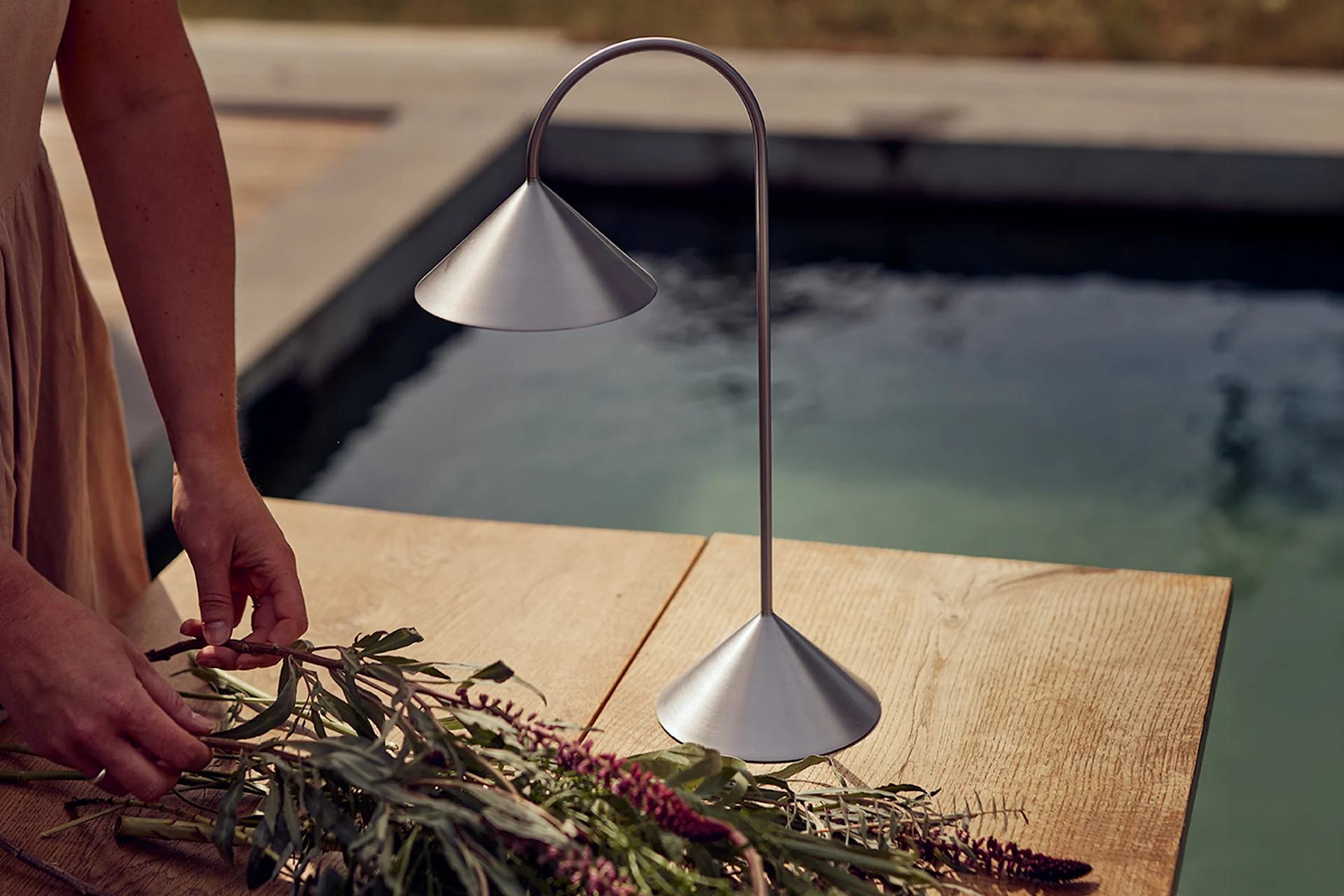 Händer arrangerar blommor på ett träbord bredvid en bordslampa i borstad metall vid en pool.