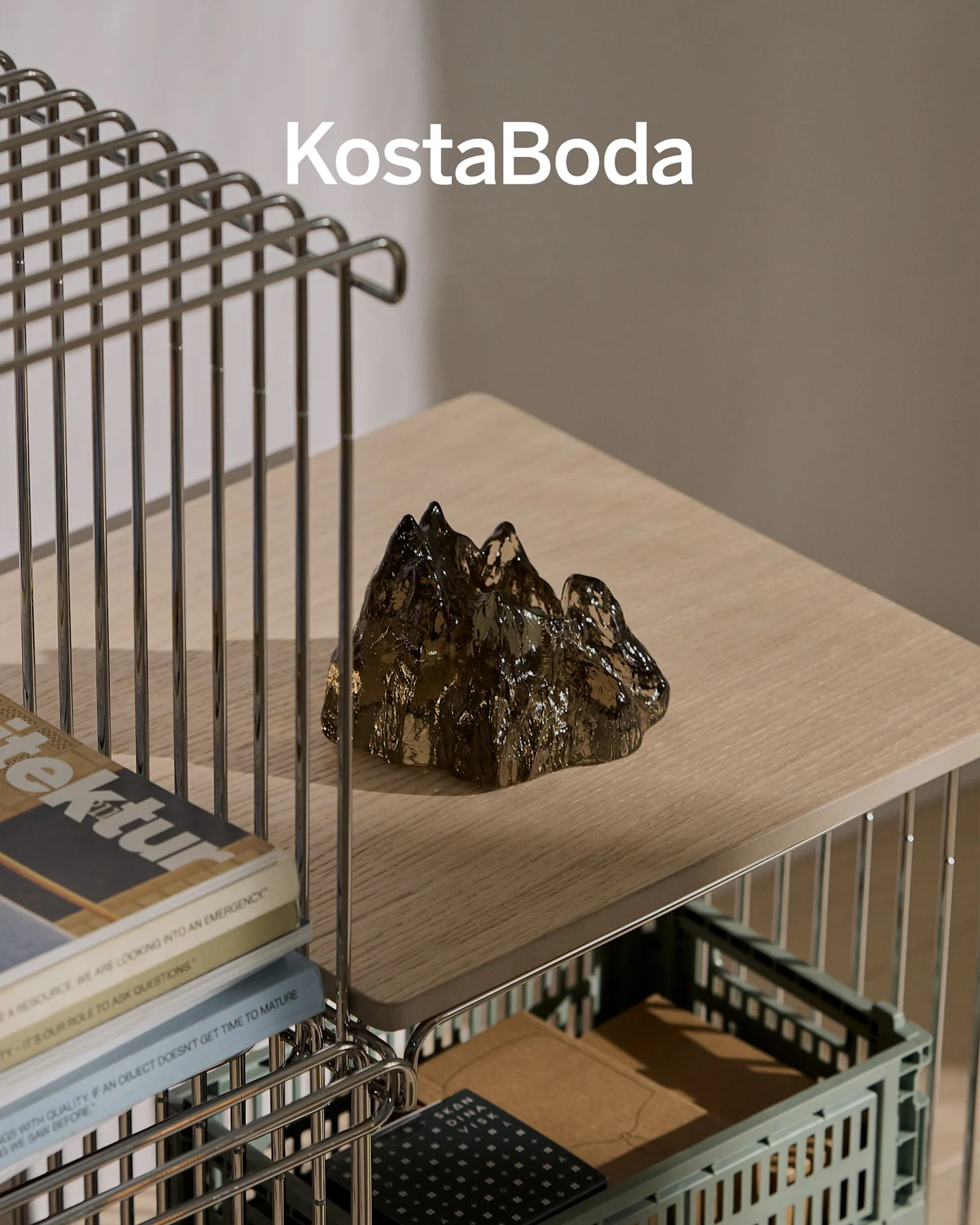 KostaBoda varumärke ovanför en mörk, transparent, bergformad glasskulptur på en ljus trähylla.
