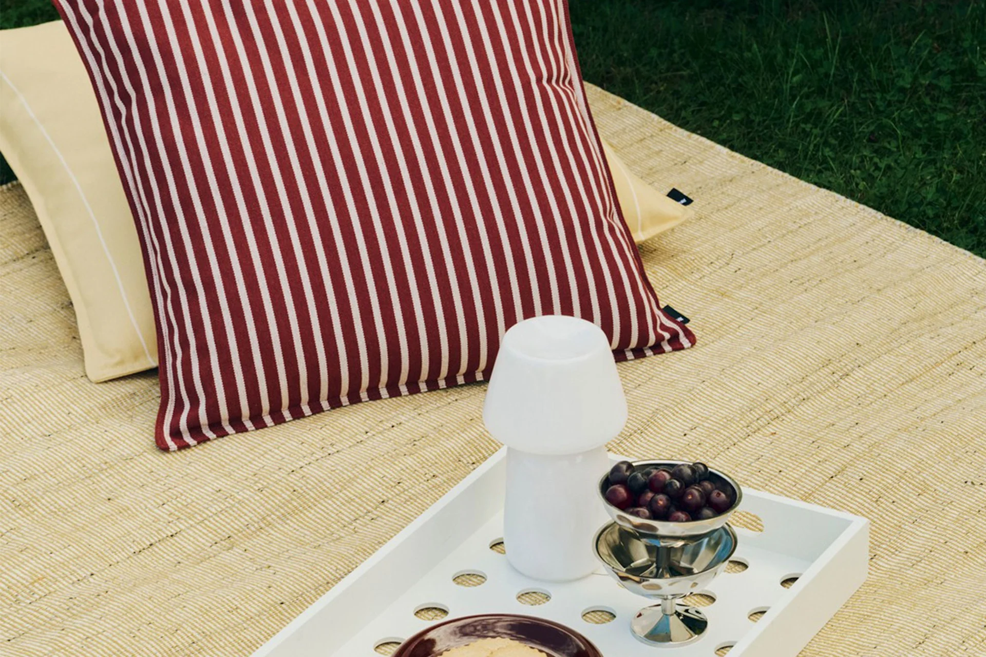 Utomhus picknickuppsättning med randig röd och vit kudde, gul kudde, vit bricka med svamplampa och en skål med bär.