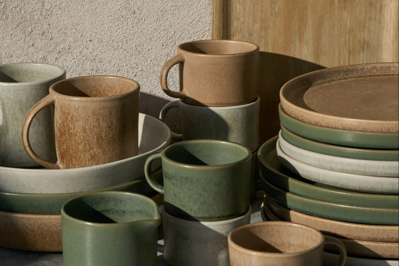 Staplade keramiska muggar, skålar och tallrikar i beige, ljusgrått och salviagrönt, med rustik finish.