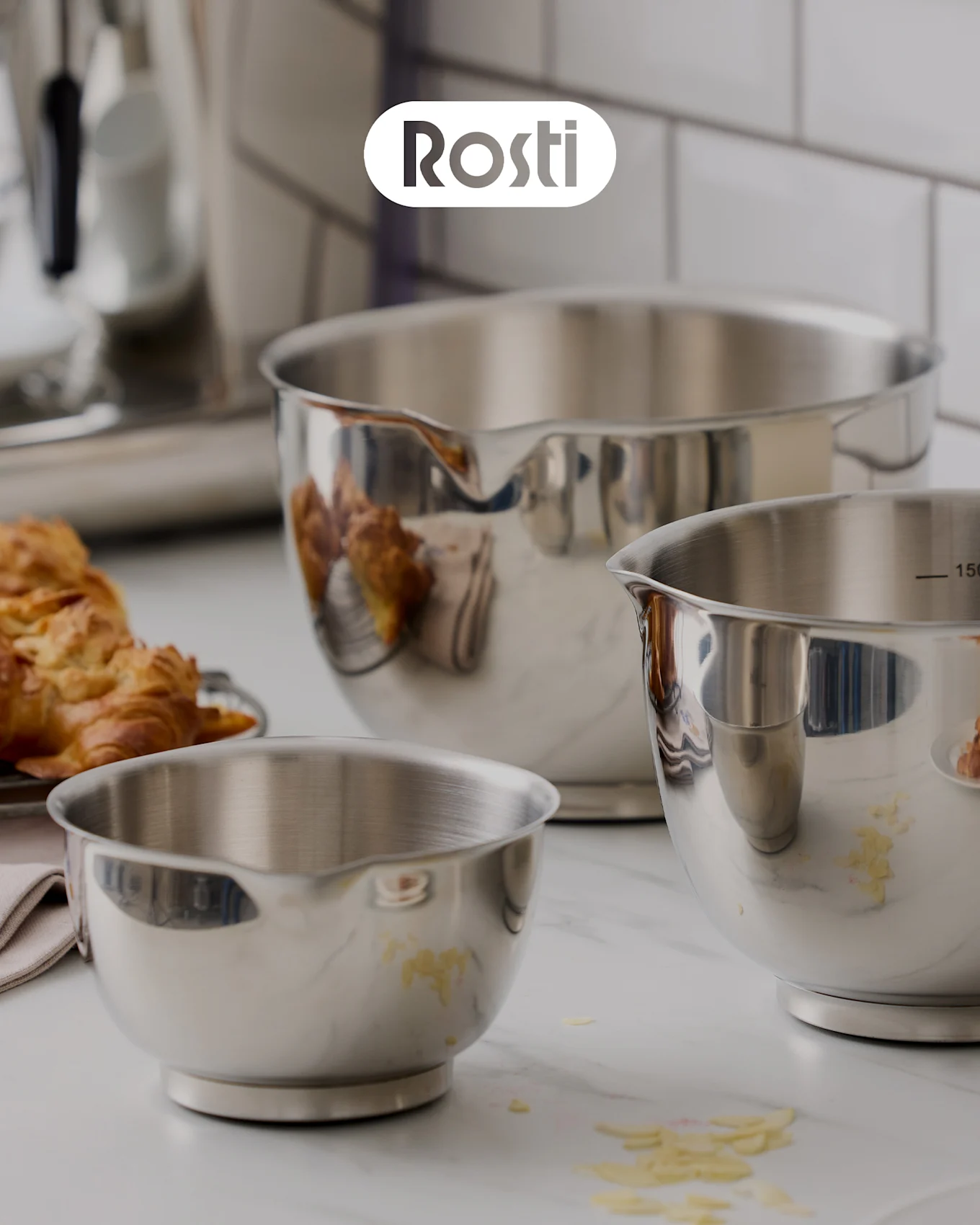 Tre Rosti-skålar på en bänk: en beige skål med en blå staplad ovanpå, båda med lock, och en liten gul skål fylld med torkade aprikoser.
