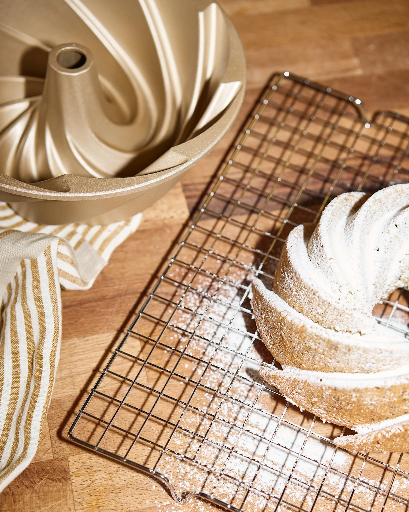 En gyllene bundt-kakform, ett galler med en pudrad bundt-kaka och en randig handduk på en träyta.