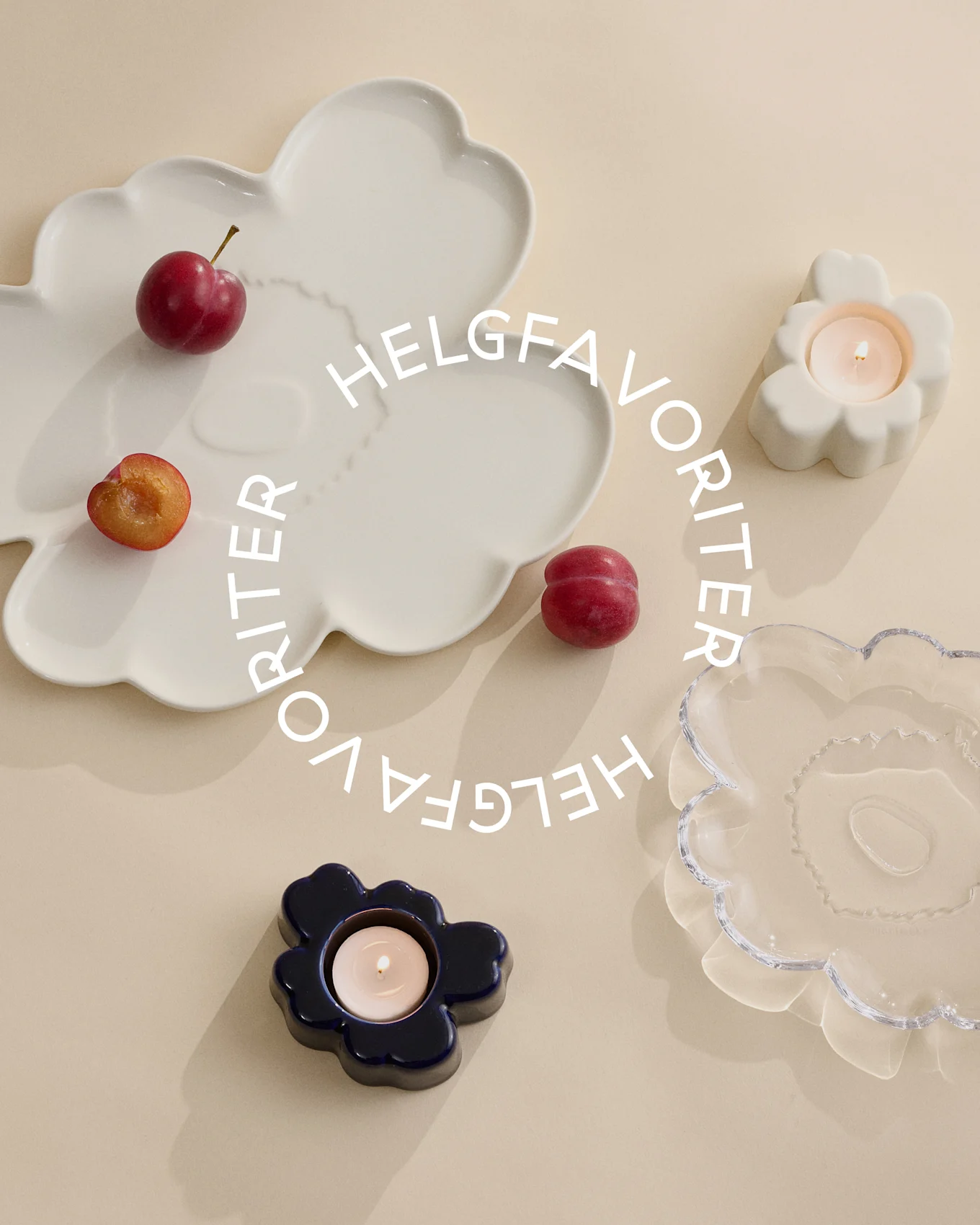 Olika blomformade fat och ljushållare i vitt, glas och mörkblått, med tända ljus och plommon. Text: Helgfavoriter.