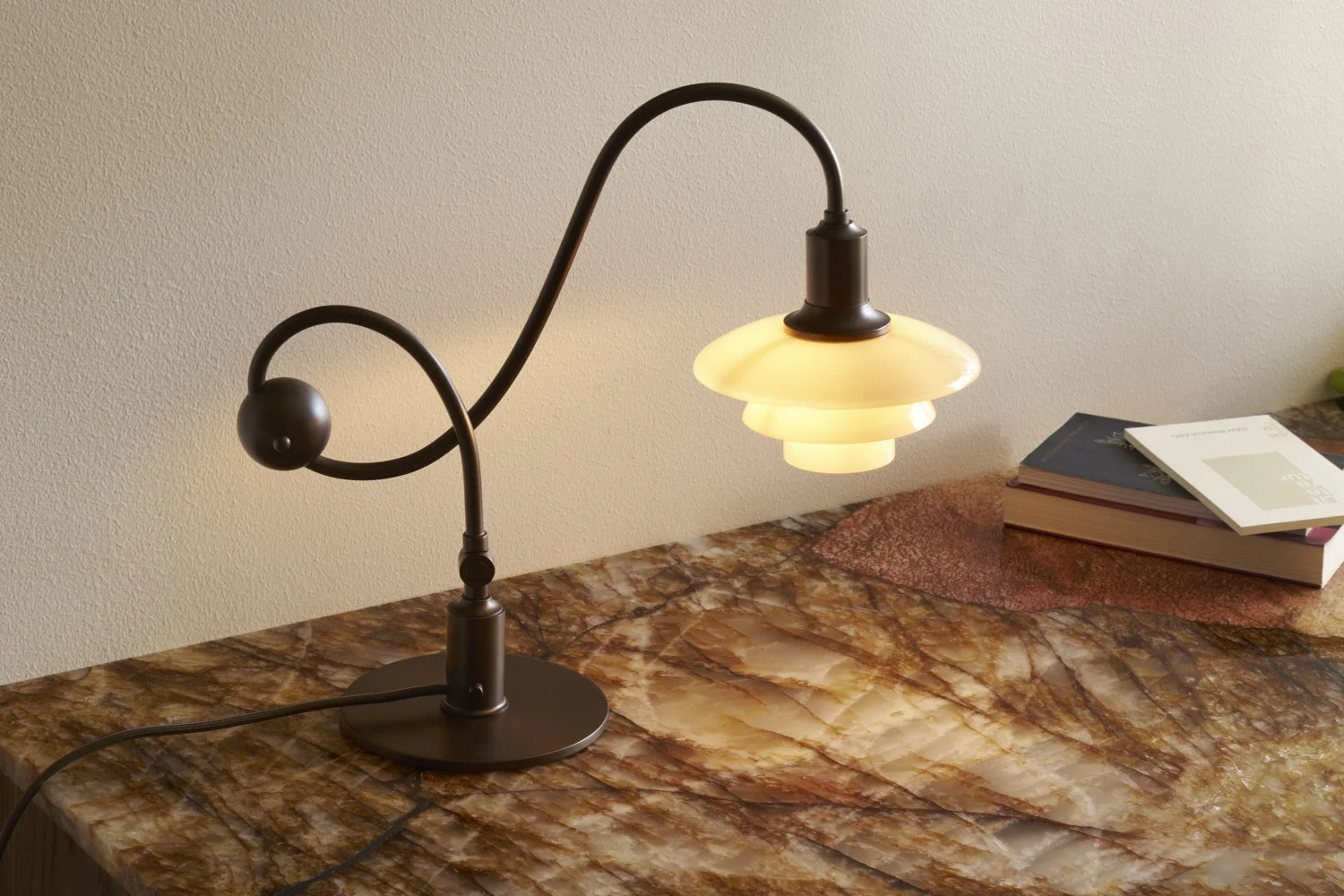 Mörk bordslampa i metall med böjd stång och skiktad, glödande vit skärm på en marmorerad yta med böcker.