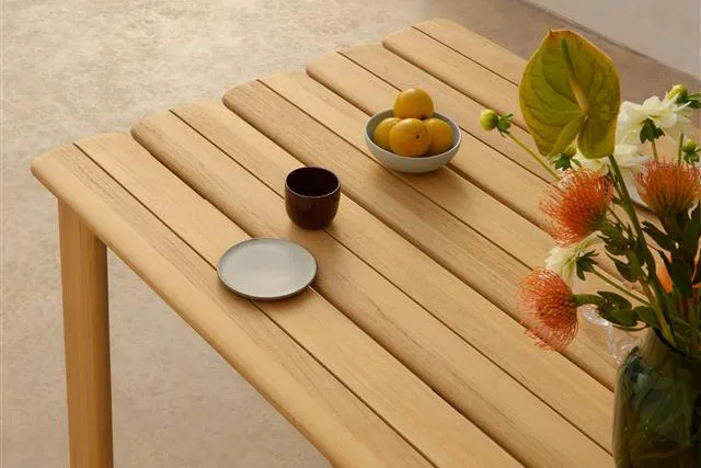 Ljust träribbsbord med en skål citroner, mörk mugg, liten tallrik och en vas med orange och vita blommor.