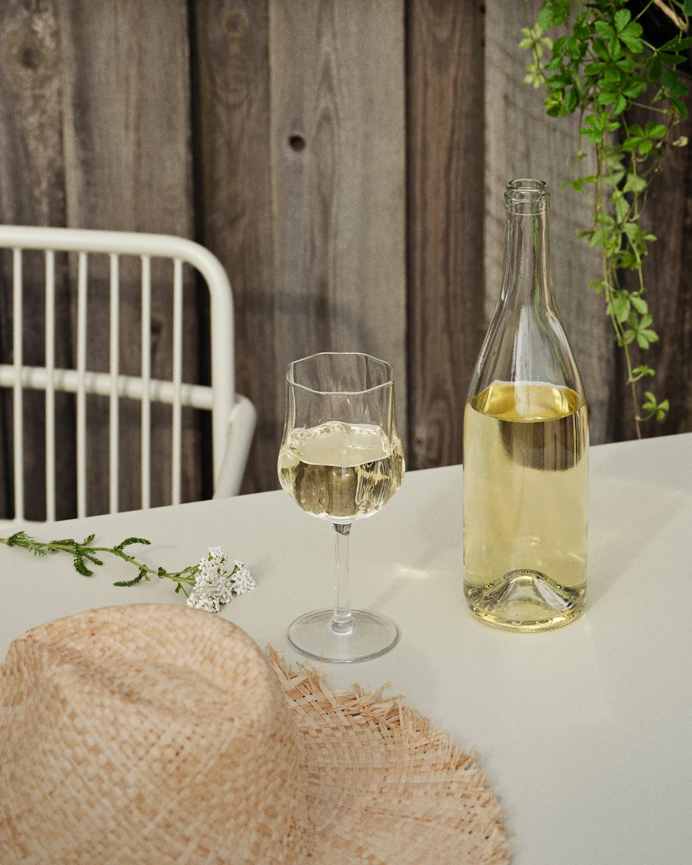 En flaska och ett glas vitt vin på ett bord med en stråhatt och vita blommor, mot en trävägg och gröna rankor.