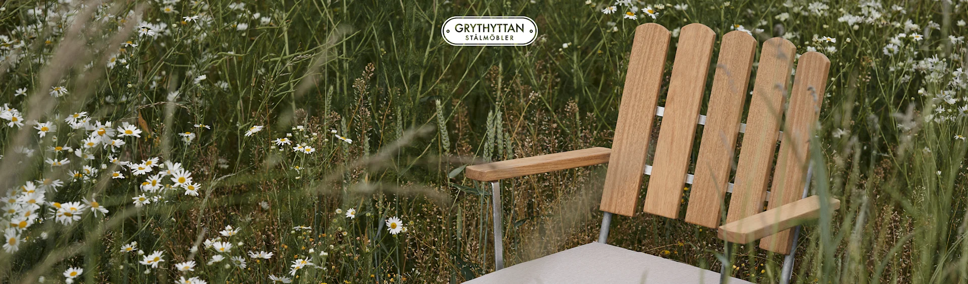 Trästol med ljus dyna i ett fält av vita prästkragar. Grythyttan Stålmöbler logotyp.