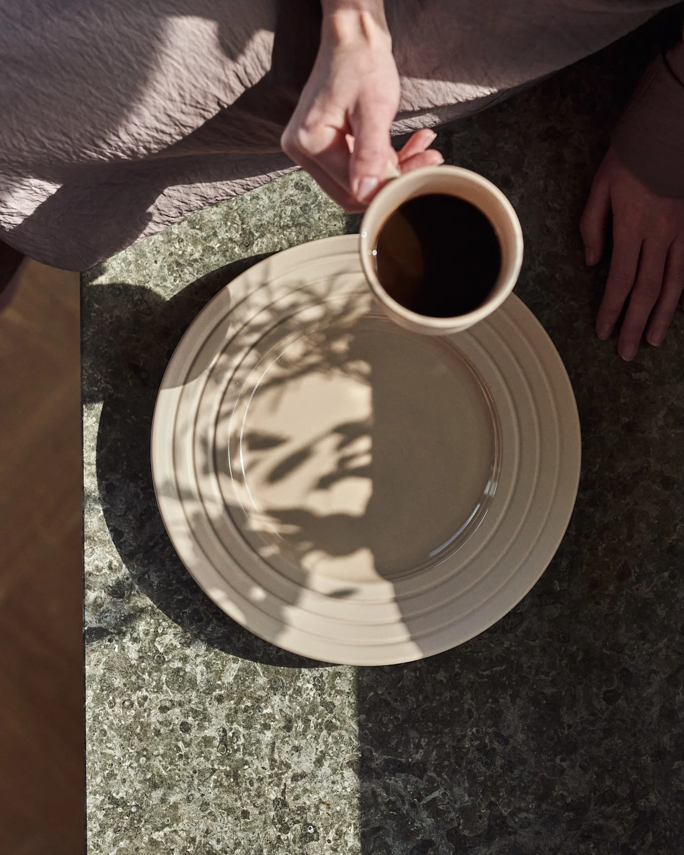 Uppifrån: händer, en håller en beige mugg med svart kaffe över en beige tallrik på ett mörkt stenbord, med bladskuggor.