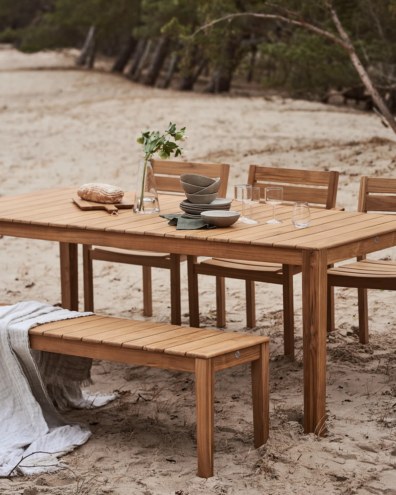 Utomhus matgrupp i trä med bord och bänk på en sandstrand. Bordet är dukat med porslin, glas, bröd och en vas med blommor.