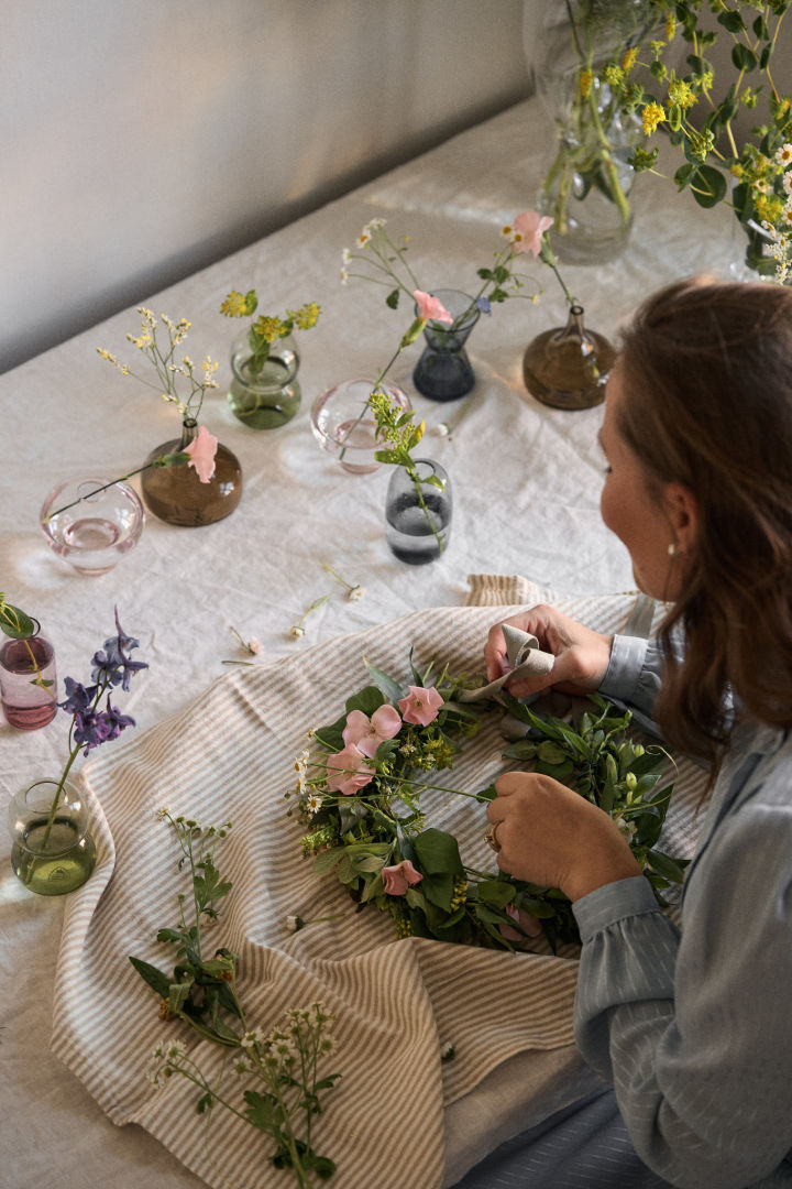 En kvinna knyter ihop en midsommarkrans omgiven av minivaser från Orrefors fyllda med vilda blommor. 