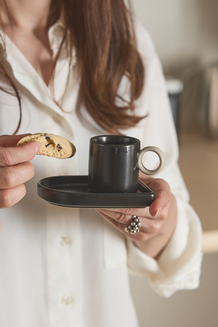 Här ser du en kvinna som håller espressokoppen Niki från Byon i ena handen och en biscotti i den andra.