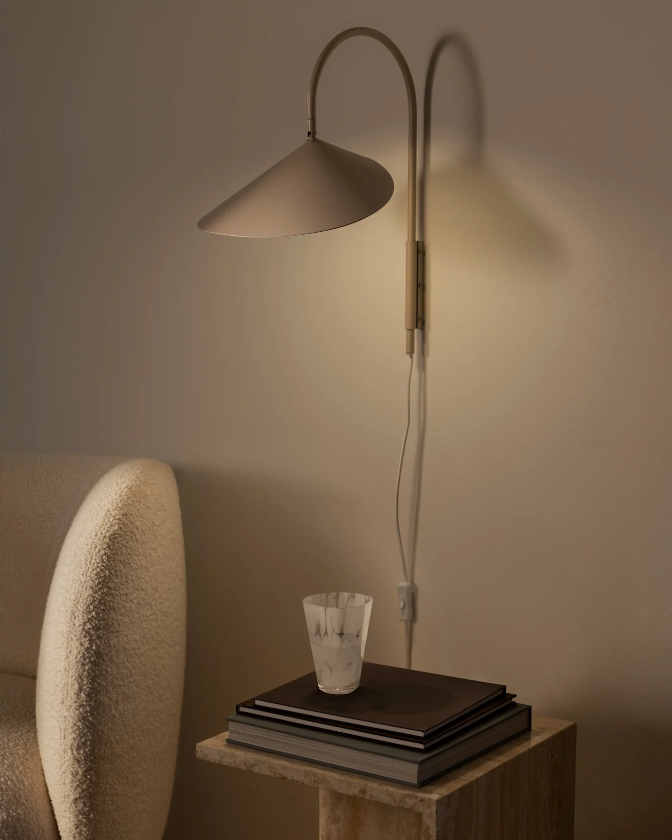 Varmt upplyst vägglampa med konisk skärm över en hög böcker och ett texturerat glas på ett stenbord, bredvid en beige fåtölj.