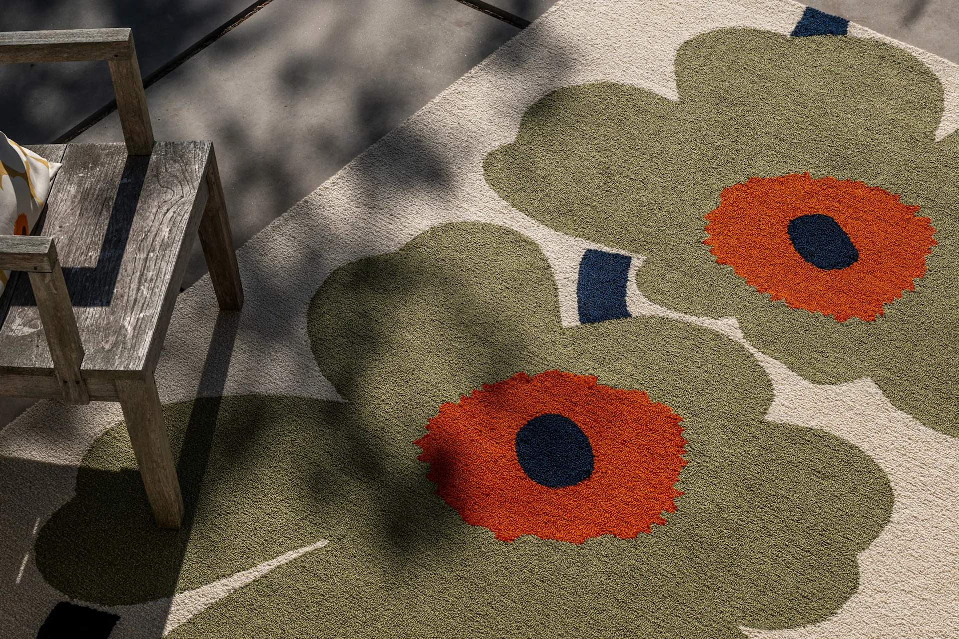 Utomhusmatta med stort olivgrönt, orange och mörkblått blommönster, intill en rustik trästol i spräckligt solljus.