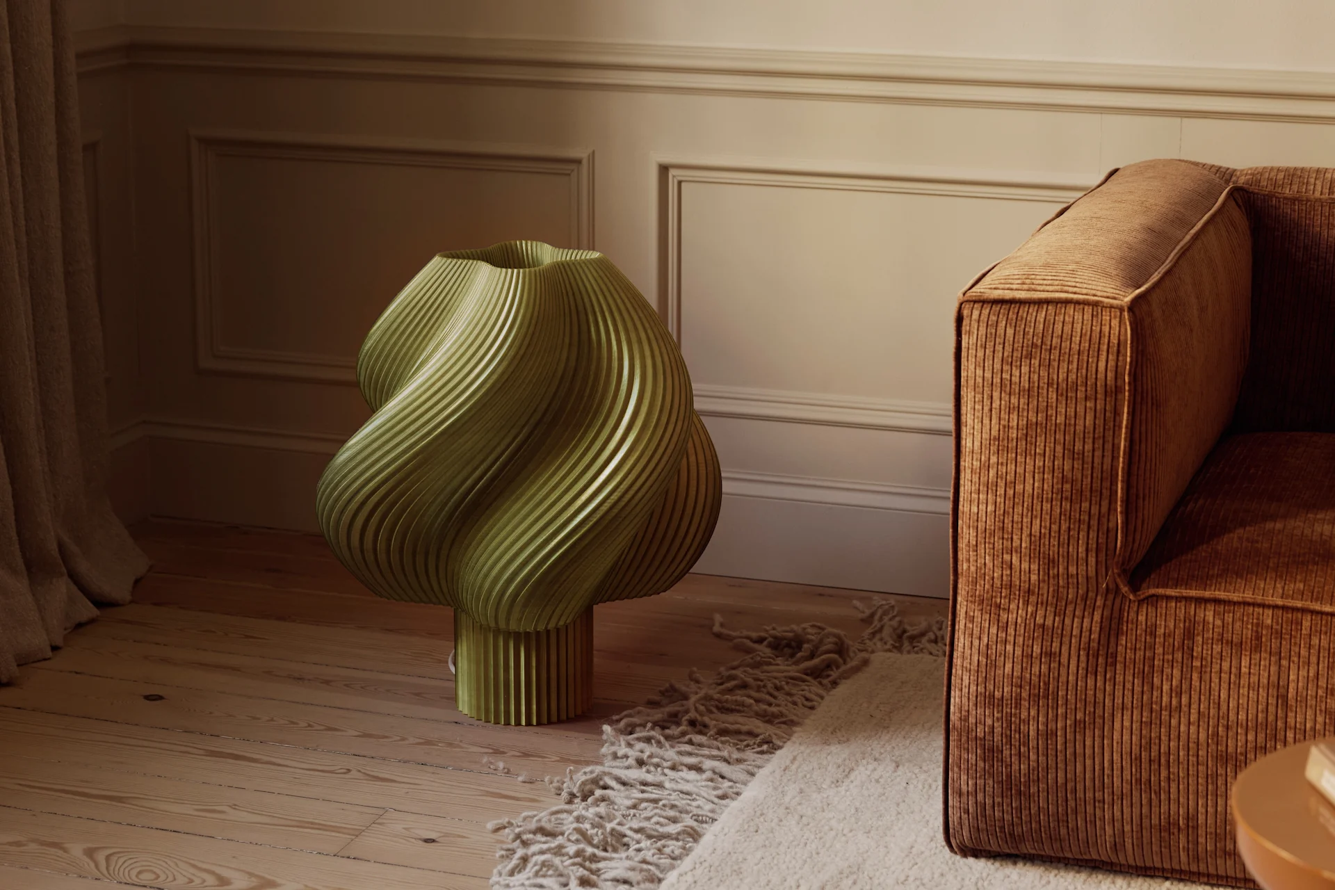 Ett olivgrönt, skulpturalt, räfflat föremål på ett trägolv bredvid en brun manchestersoffa och en texturerad matta i ett rum med profilerade väggar.