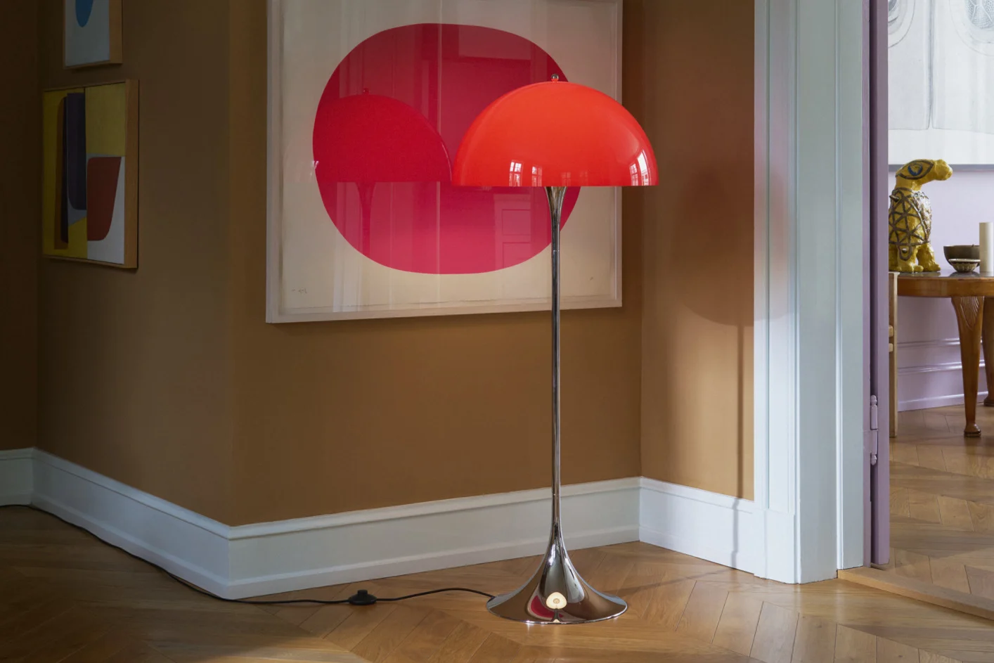 En klar röd kupolformad golvlampa med kromstativ står på ett trägolv i ett rum med bruna väggar och modern konst.